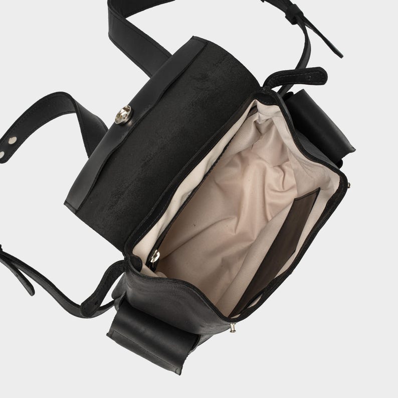 interior view of black European leather backpack with flap open, showing beige waterproof lining, internal zip pocket, and slip pocket for organization