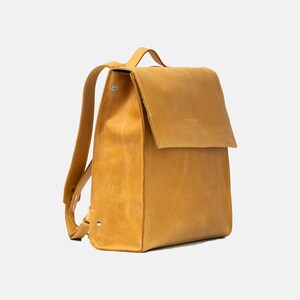May include: Mustard yellow leather backpack with a flap closure. The bag has adjustable shoulder straps and a top handle. The leather has a textured appearance. The backpack is rectangular with rounded corners.