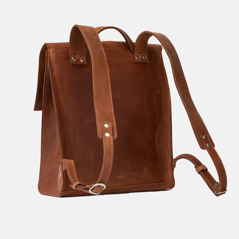 May include: A brown leather backpack with adjustable shoulder straps and a flap closure. The bag features a rectangular shape with a top handle and silver-toned hardware. The leather has a textured appearance.