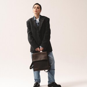 May include: A dark brown leather backpack with adjustable straps is held in front of a person wearing a dark gray blazer, a denim shirt, and blue jeans. The person is also wearing black shoes.