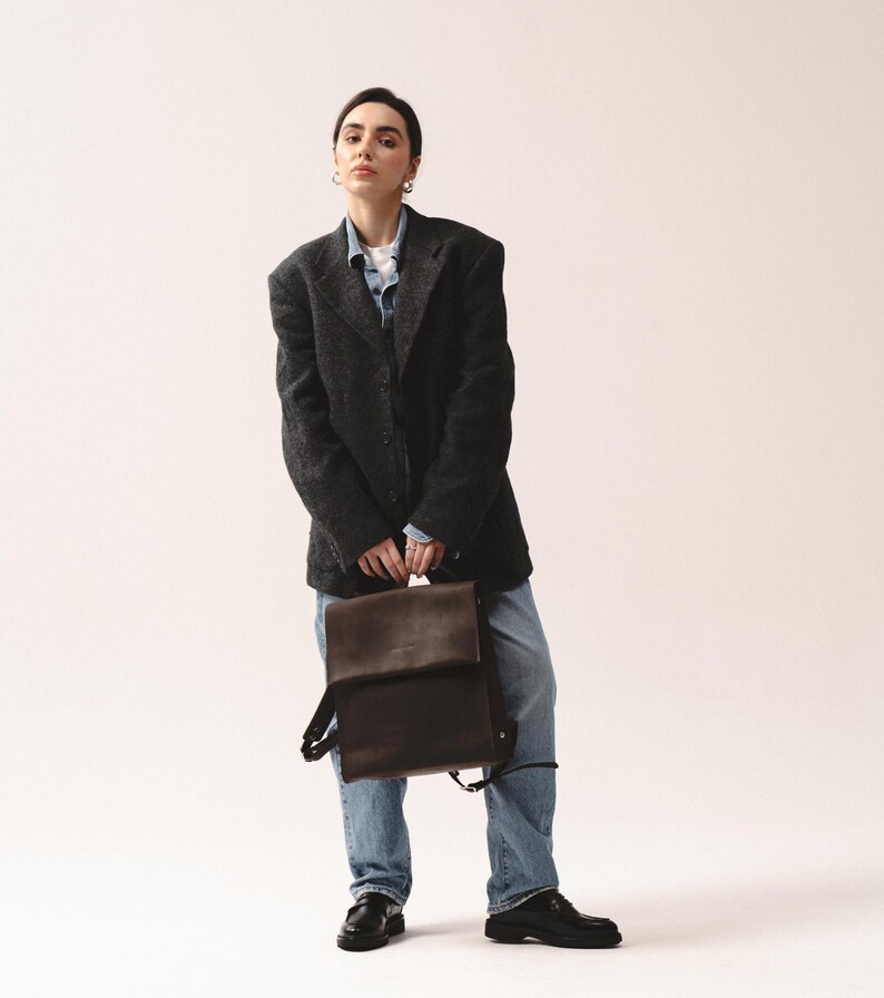May include: A dark brown leather backpack with adjustable straps is held in front of a person wearing a dark gray blazer, a denim shirt, and blue jeans. The person is also wearing black shoes.