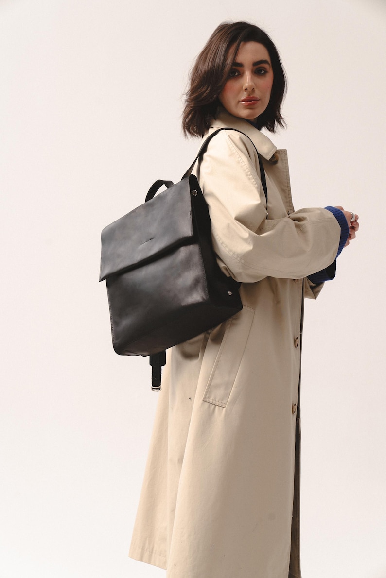 May include: A black leather backpack with a flap closure. The backpack has adjustable straps and a top handle. The person is wearing a tan trench coat with a blue sweater underneath.