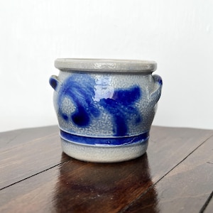 May include: A small, gray and blue ceramic pot with two small handles. The pot features a blue floral design and a blue stripe around the base. The rim is a lighter gray color. The pot is sitting on a dark wooden surface.