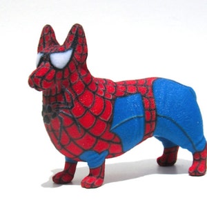May include: A 3D printed figurine of a dog dressed as Spiderman. The dog is red and blue with a black spider web design. The dog has white eyes and a black spider on its chest.