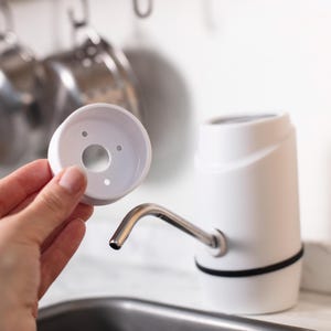 May include: White plastic water bottle pump with a silver spout and a white plastic base. The pump is detached from the base and is being held in a hand.