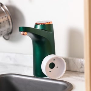 May include: A green and copper water dispenser pump with a white plastic base. The pump is sitting on a white countertop next to a stainless steel sink.
