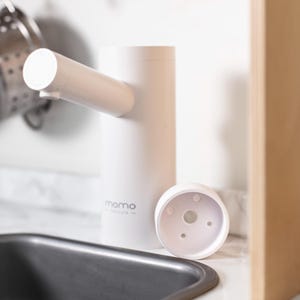 May include: White electric soap dispenser with a pump and a round base. The dispenser is labelled "momo"