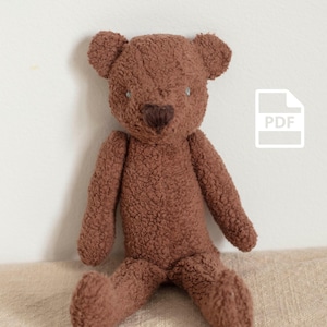 Op de afbeelding: Een bruine teddybeer met een getextureerde, pluche buitenkant. De beer heeft kleine, ronde oren, een hartvormige neus en lichtblauwe ogen. Hij zit op een beige ondergrond tegen een witte achtergrond. Een PDF-pictogram staat op de achtergrond.
