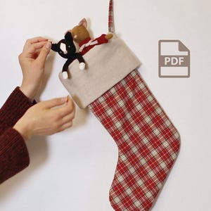 Può includere: Una calza di Natale con un motivo a quadri rossi e bianchi, riempita con un peluche di gatto bianco e nero e un peluche di topo marrone. La calza ha un polsino beige e un anello a righe rosse e bianche per appenderla.