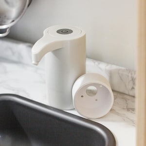 May include: A white, automatic soap dispenser with a curved spout and a detached base. The dispenser has a round top with a power button. The dispenser is on a marble-patterned countertop near a black sink.