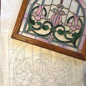 May include: A stained glass window design featuring a floral motif with pink, green, and blue glass pieces. The design is framed in wood and is accompanied by a paper pattern.