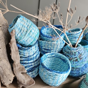 Może przedstawiać: Kolekcja małych, wiklinowych koszyków w odcieniach niebieskiego i białego. Koszyki są wykonane z naturalnych materiałów i mają rustykalny, ręcznie robiony wygląd.