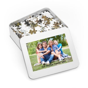 May include: A white metal tin with a photo of a family of four sitting on a blanket in a grassy area. The photo is captioned "The Fletcher Family 2021". The tin is filled with white jigsaw puzzle pieces.