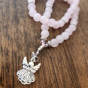 May include: A pink rose quartz beaded necklace with a silver angel pendant. The necklace features round, light pink beads and silver accents. The angel pendant is detailed with wings and a halo. The necklace is displayed on a wooden surface.
