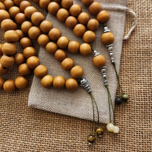 May include: Three strands of wooden beads with silver accents and green tassels. The beads are a light brown color and are arranged in a circle. The beads are on a brown burlap surface.