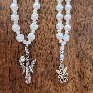 May include: Two white beaded rosaries with angel pendants. The beads are shaped like roses. One rosary has a larger angel pendant with a crystal accent. The other has a smaller angel pendant.