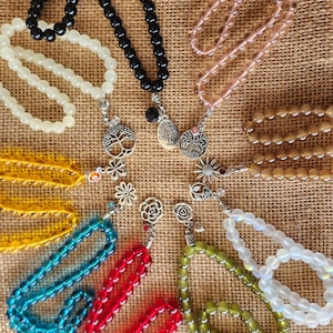 May include: Assortment of colorful beaded prayer necklaces with silver charms. The charms include a tree of life, a flower, and a heart. The necklaces are arranged in a circle on a burlap background.