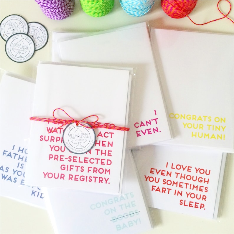 May include: A set of greeting cards with humorous messages for a new baby. The cards are white with black text and are packaged in clear plastic sleeves. One card has a pink and red ribbon tied around it with a small round tag that says "SPADE STATIONERY ETSY.COM".
