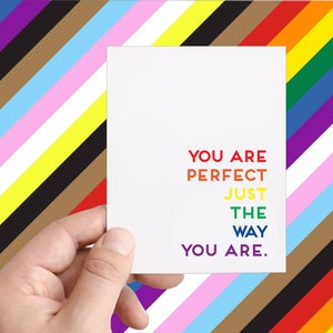 May include: A white card with the text "You are perfect just the way you are." in rainbow colors. The card is held by a hand against a background of colorful stripes.