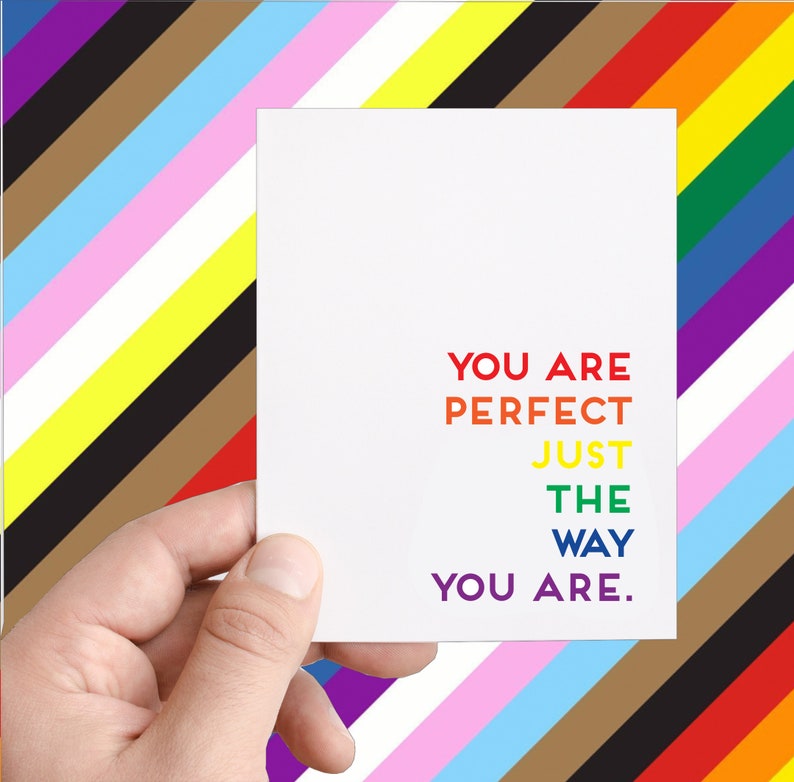 May include: A white card with the text "You are perfect just the way you are." in rainbow colors. The card is held by a hand against a background of colorful stripes.