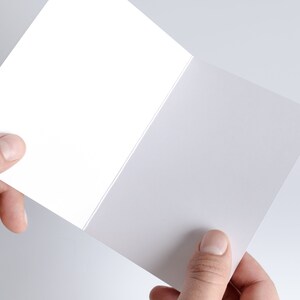 May include: A blank white card folded in half, held by a hand. The card is a standard size, with a smooth, matte finish.