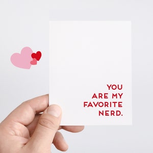 Pu&ograve; includere: Un biglietto bianco con testo rosso che dice "Sei il mio nerd preferito." Due cuori rosa e un cuore rosso sono nell'angolo in alto a sinistra del biglietto.