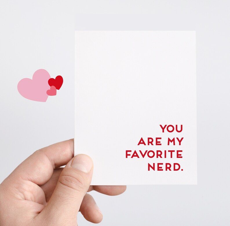 Pu&ograve; includere: Un biglietto bianco con testo rosso che dice "Sei il mio nerd preferito." Due cuori rosa e un cuore rosso sono nell'angolo in alto a sinistra del biglietto.