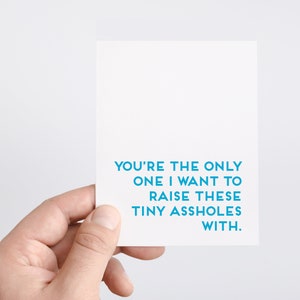 May include: A white card with teal blue text that reads "YOU'RE THE ONLY ONE I WANT TO RAISE THESE TINY ASSHOLES WITH". The card is held by a hand against a white backdrop.