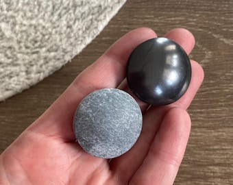 May include: Two circular objects, one with a smooth, polished, dark charcoal-colored surface that reflects light, and the other with a matte, textured, light gray surface showing natural stone-like variations in its circular form.