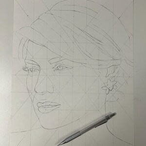 May include: A graphite pencil sketch on white paper featuring a portrait of a person wearing a hat and a floral earring. The drawing includes geometric grid lines for proportion and a mechanical pencil resting across the bottom right corner.