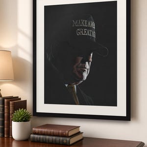 May include: A framed portrait print featuring a dark, moody side profile of a person wearing a dark baseball cap with the text MAKE AMERICA GREAT AGAIN embroidered in white. The subject wears a dark suit jacket and a gold-toned necktie.