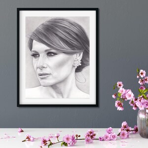 May include: A black and white charcoal portrait drawing of a person with side-swept hair and a floral earring, encased in a thin black rectangular frame. The artwork is displayed on a dark gray wall above a white surface with pink flowers.