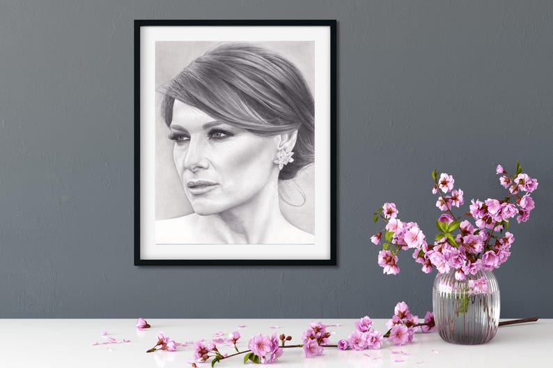 May include: A black and white charcoal portrait drawing of a person with side-swept hair and a floral earring, encased in a thin black rectangular frame. The artwork is displayed on a dark gray wall above a white surface with pink flowers.