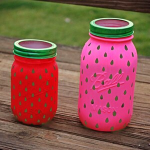 May include: Two glass jars painted to look like strawberries. The larger jar is pink with green accents and the smaller jar is red with green accents. Both jars have a green lid.