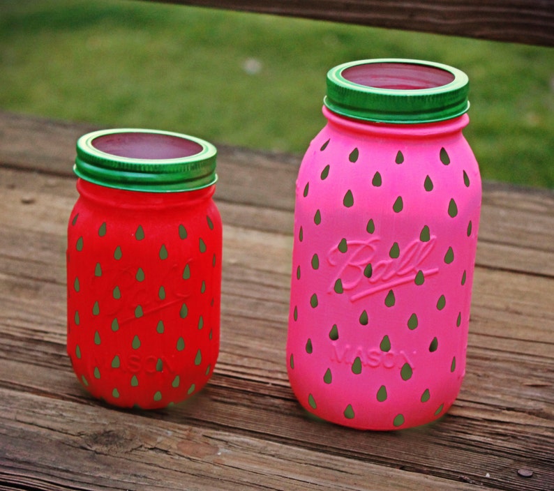 May include: Two glass jars painted to look like strawberries. The larger jar is pink with green accents and the smaller jar is red with green accents. Both jars have a green lid.
