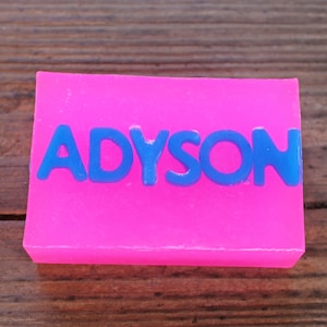 May include: A bright pink rectangular soap bar with the name "ADYSON" in blue letters across the top. The soap is set against a wooden background, creating a contrast between the vibrant colors and the natural wood.