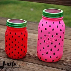 May include: Two glass jars painted to look like watermelons. The larger jar is pink with black watermelon seeds and the smaller jar is red with black watermelon seeds. Both jars have green lids.
