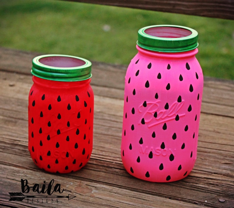 May include: Two glass jars painted to look like watermelons. The larger jar is pink with black watermelon seeds and the smaller jar is red with black watermelon seeds. Both jars have green lids.