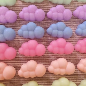 May include: A collection of pastel-coloured cloud-shaped wax melts in various shades of green, purple, blue, pink, orange, and yellow. The wax melts are arranged in rows on a brown woven surface.