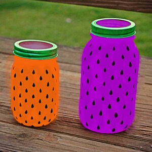 May include: Two glass jars painted to look like watermelons. The smaller jar is orange with black seeds and the larger jar is purple with black seeds.