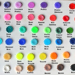 K&ouml;nnte beinhalten: Eine Karte mit 40 kleinen, blumenf&ouml;rmigen Kn&ouml;pfen in verschiedenen Farben, darunter Lila, Rosa, Rot, T&uuml;rkis, Gr&uuml;n, Blau, Gelb, Orange, Wei&szlig;, Schwarz, Silber, Gold, Kupfer und Bronze. Jeder Knopf ist mit seinem Farbnamen beschriftet.