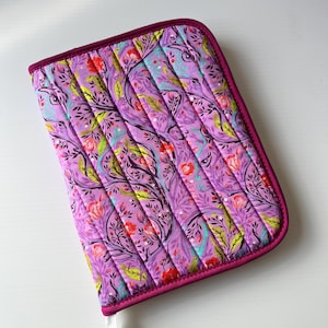 May include: A padded case with a floral design in purple, red, and green. It has a dark pink border and vertical quilting. The pattern features stylized flowers and branches. The case is quilted.