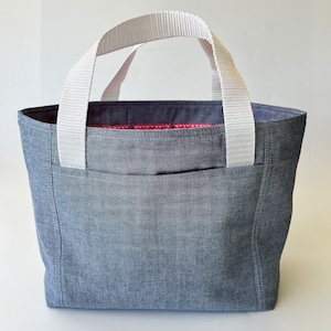 May include: A grey tote bag with white handles and a front pocket. The bag has a rectangular shape and a patterned red and blue interior lining. The handles are made of a sturdy, woven material.