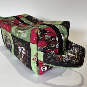 May include: A green, red and black zippered travel bag with a black handle. The bag is decorated with a repeating pattern of characters from the film "The Nightmare Before Christmas". The text "Scaring Twice as Nice" is printed on the handle.