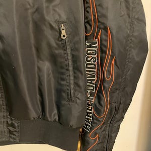 May include: Black satin jacket with a zippered pocket and an orange and white flame design with the text "Harley-Davidson" on the right side.
