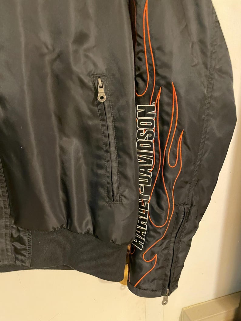May include: Black satin jacket with a zippered pocket and an orange and white flame design with the text "Harley-Davidson" on the right side.