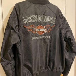 May include: Black bomber jacket with a Harley-Davidson logo and flames on the back. The logo is white with red accents and says "Harley-Davidson Motorcycles".
