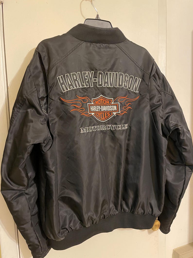 May include: Black bomber jacket with a Harley-Davidson logo and flames on the back. The logo is white with red accents and says "Harley-Davidson Motorcycles".