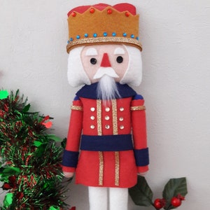 May include: A felt nutcracker doll wearing a red and gold uniform with a blue sash and a gold crown with blue jewels. The doll has a white beard and white hair. The doll is standing on a wooden surface.
