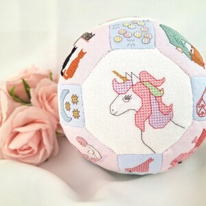May include: A soft, pink, multi-faceted ball with embroidered panels. The panels feature various designs, including a unicorn, cats, hearts, and celestial elements. The ball is surrounded by pink roses.